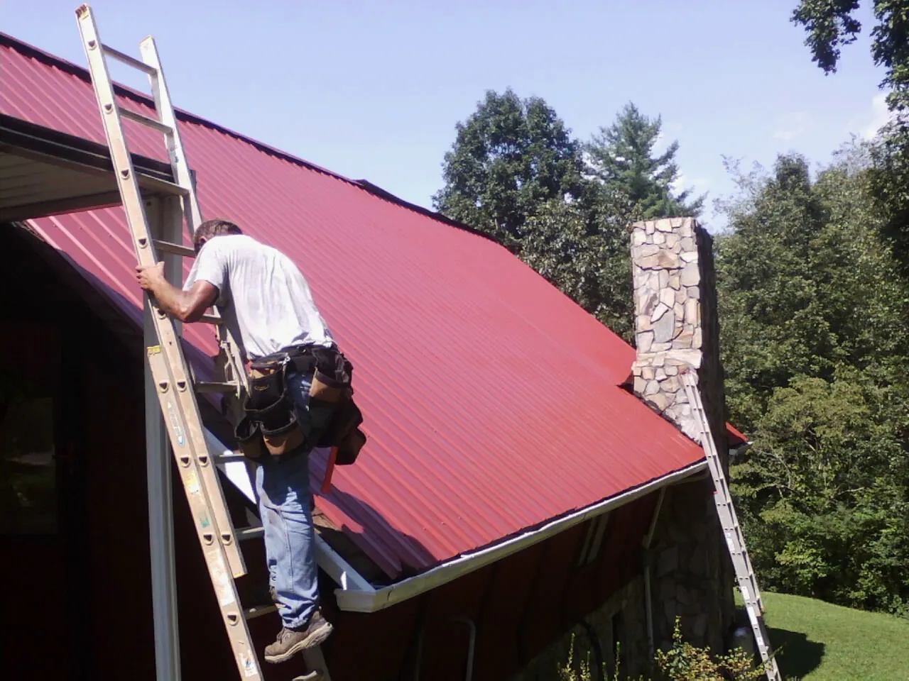 Roofer on a ladder beside a red metal roof and stone chimney — Soffit & Fascia Repair by Roofing Brothers in Kensington